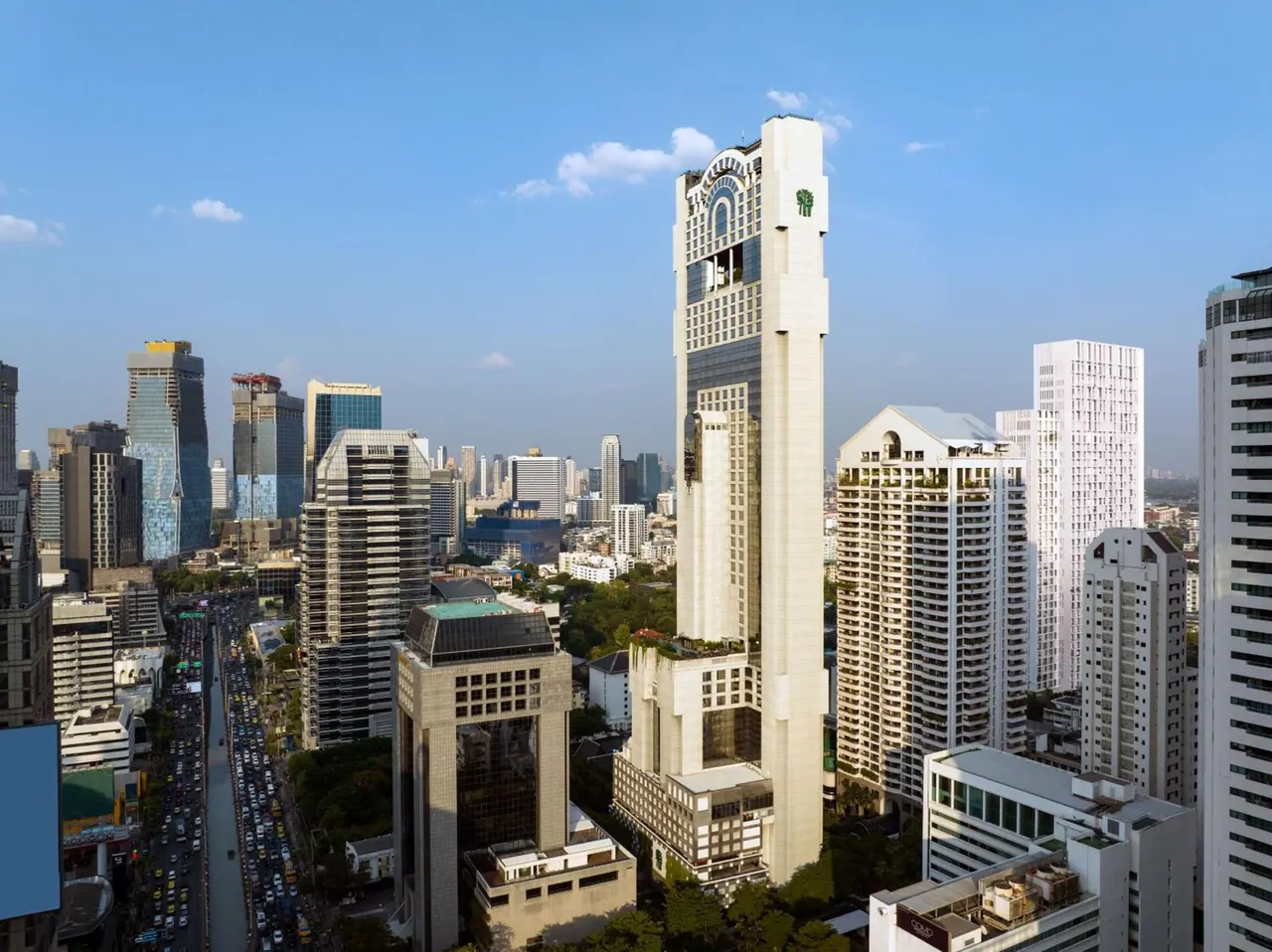 Aerial view of a tall cream-coloured tower with a green logo rising above surrounding high-rise buildings and a busy road full of traffic.