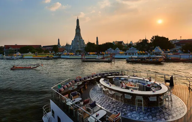 A circular outdoor bar on a deck has high stools and tables overlooking the Chao Phraya river and Wat Arun at sunset.
