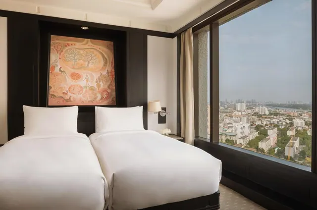 A bedroom with two white beds, a large painting on a dark wall, and a floor-to-ceiling window looking over a city with a river in the distance.