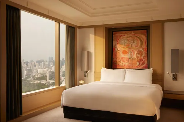 A bedroom with a king bed, a large colourful painting on the wall and floor-to-ceiling windows overlooking a hazy city skyline.