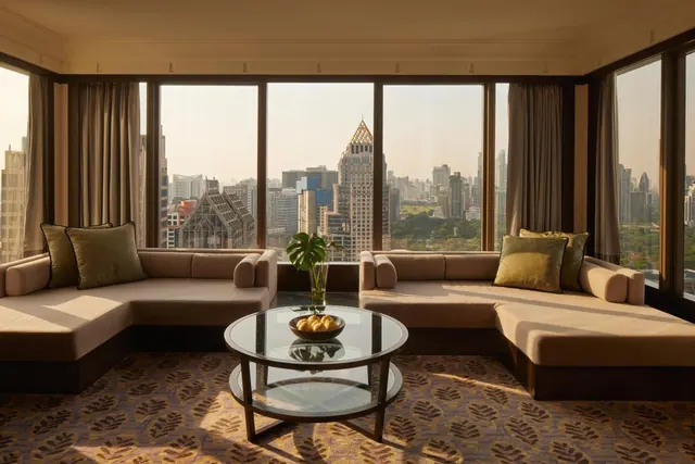A living area with two low sofas and a round glass coffee table faces large windows overlooking a city skyline and a green park.