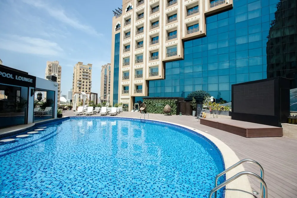 An outdoor pool with blue mosaic tiles, white sun loungers on the deck, and a glass-fronted hotel tower and residential buildings behind it.
