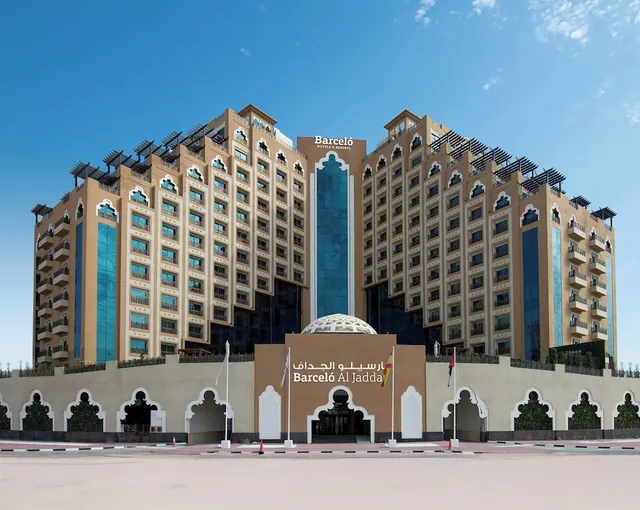 The front exterior of the Barceló Al Jadda hotel building under a clear blue sky, with flags and an arched entrance at street level.