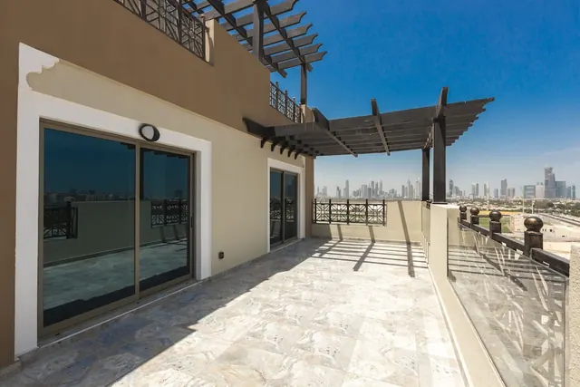A bare tiled rooftop terrace with a wooden pergola, glass balustrade and sliding glass doors overlooks a distant city skyline under a clear blue sky.