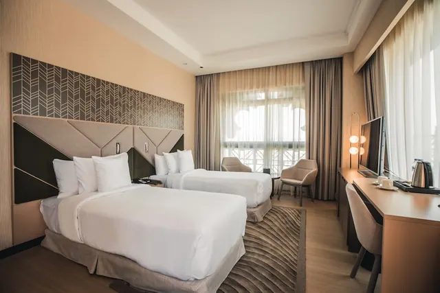 A twin bedroom with two made beds, a patterned upholstered headboard, two armchairs by a window, a work desk and a wall-mounted television.