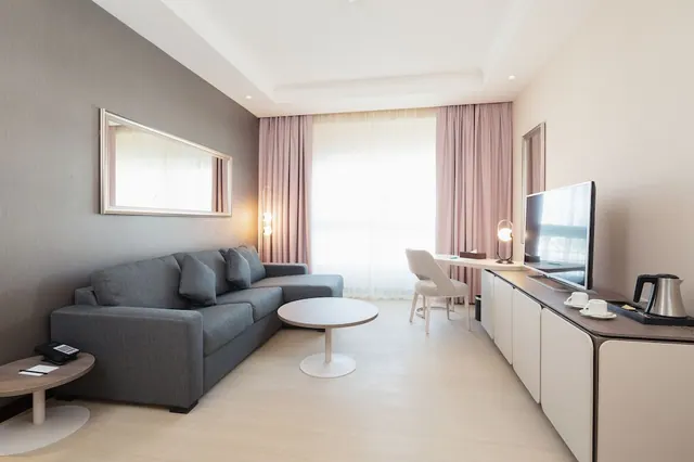 A hotel living area with a grey sofa, round coffee table, desk and chair by sheer-curtained windows, and a flat-screen TV on a sideboard with a kettle and cups.