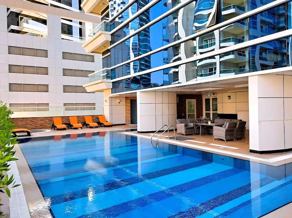 An outdoor swimming pool with blue tiled lining and orange sun loungers sits beside a covered seating area and a glass-fronted high-rise building.