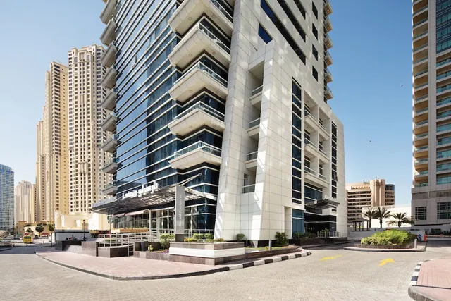 Exterior of a tall hotel tower with stacked balconies and a glass facade, with an entrance canopy and paved forecourt at street level.