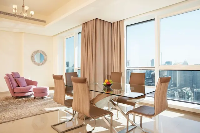 A dining area with a glass table and six upholstered chairs sits beside a pink armchair, with large windows showing a city skyline.