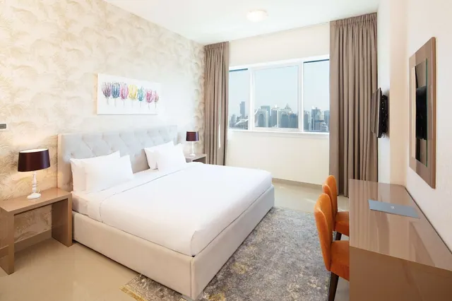 A bedroom with a made king bed, two bedside lamps, a wall-mounted television, a desk with orange chairs, and a window looking out over a city skyline.