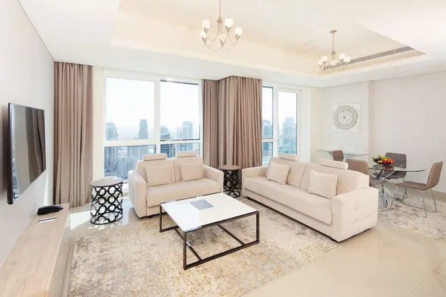 A living room with two cream sofas, a white coffee table, a wall-mounted television and floor-to-ceiling windows overlooking city towers.