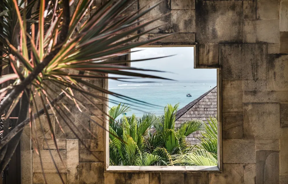 A stone wall frames a window looking out over the sea, with palm trees and a shingled roof below and a boat on the water.