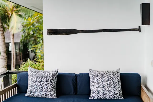A navy sofa with patterned cushions sits on a covered terrace beside a white wall with a wooden paddle, with palm trees and foliage visible outside.