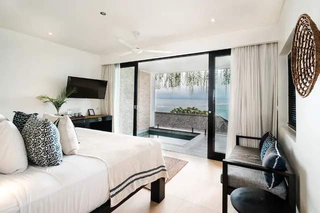 A bedroom with a made king bed, a wall-mounted television, and sliding glass doors opening onto a terrace with a small plunge pool and an ocean view.
