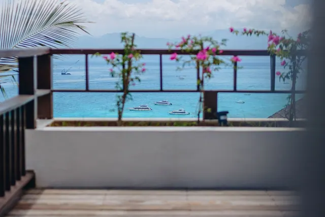 A wooden balcony railing and white wall frame a view of turquoise sea with several boats and distant mountains, with pink bougainvillea in front.
