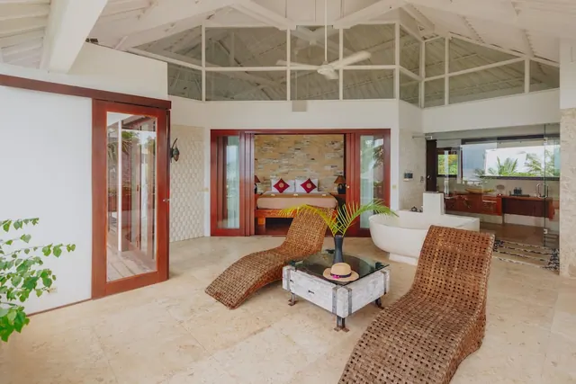 An open-plan villa room with wicker loungers, a freestanding bathtub, a bed visible through a doorway, and palm trees seen through glass.