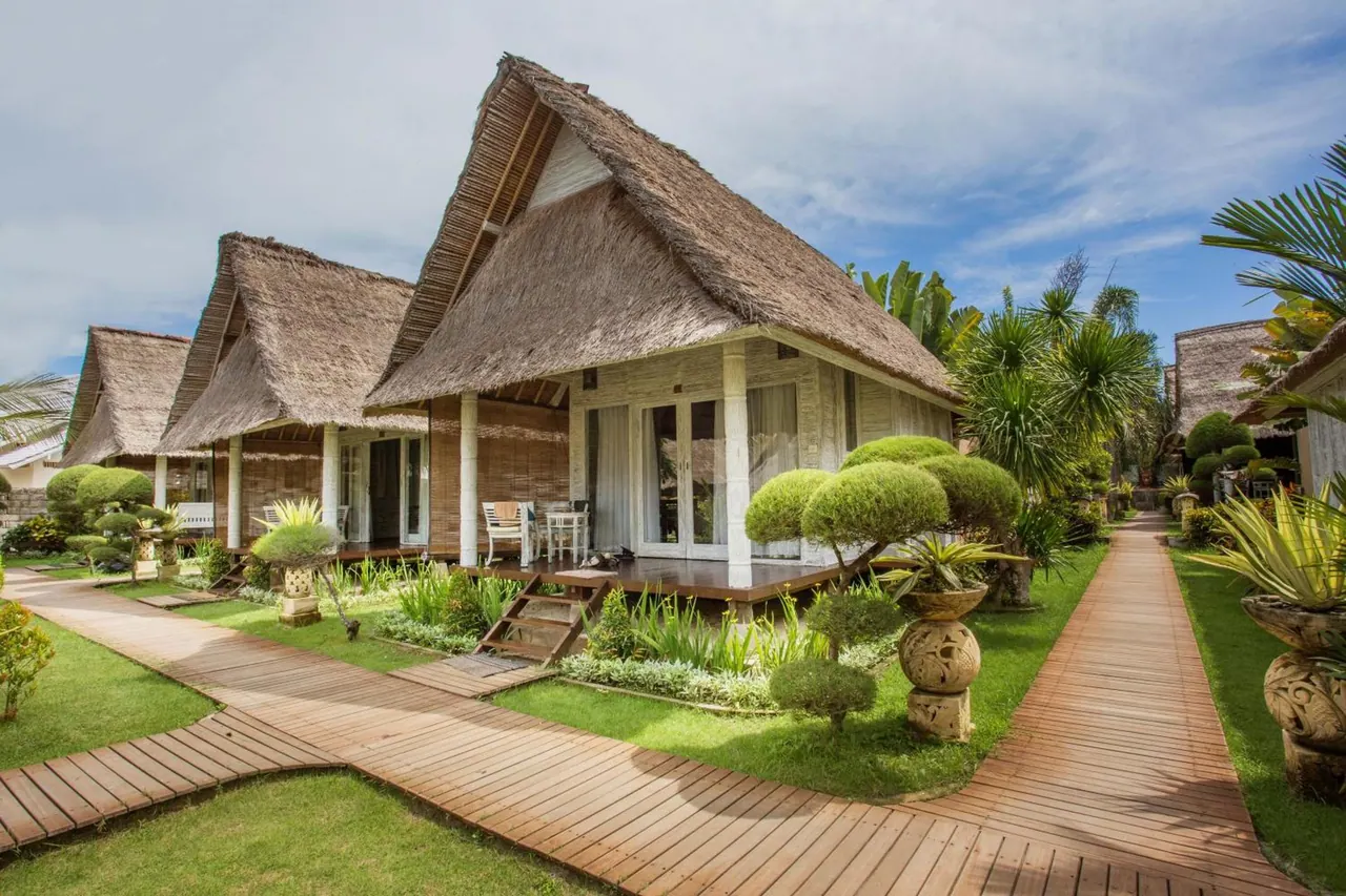 Thatched-roof bungalows with a timber deck and walkway stand among lawns and clipped topiary under a partly cloudy sky.