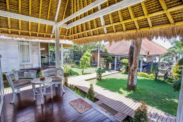 A covered thatched terrace with white wooden chairs and a small round table overlooks a lawn garden and a wooden walkway.