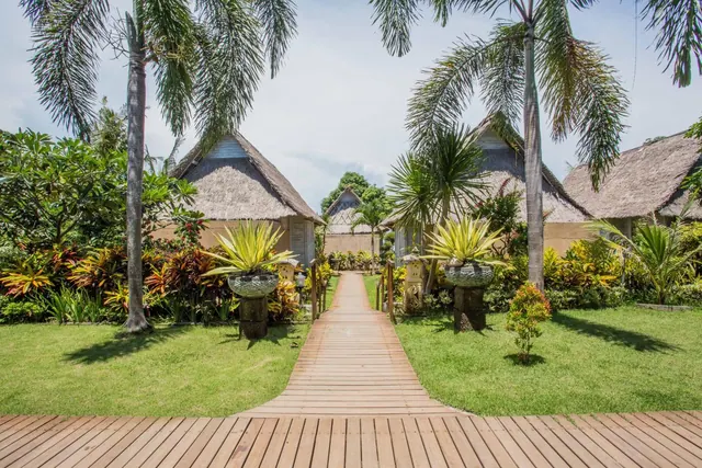 A wooden walkway leads between lawns and thatched-roof bungalows lined with palm trees and potted plants.
