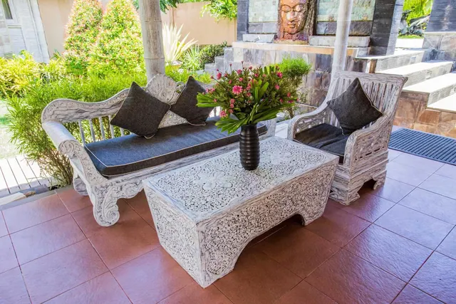 A covered terrace with a carved whitewashed wooden sofa, armchair and coffee table, a flower vase on the table, and a stone water feature with a Buddha face in the background.