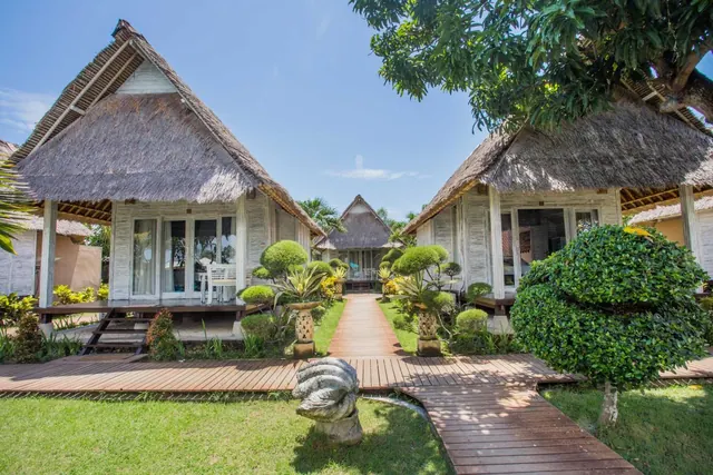 Two thatched-roof bungalows stand either side of a wooden walkway that runs through a lawn with trimmed shrubs and a stone sculpture.