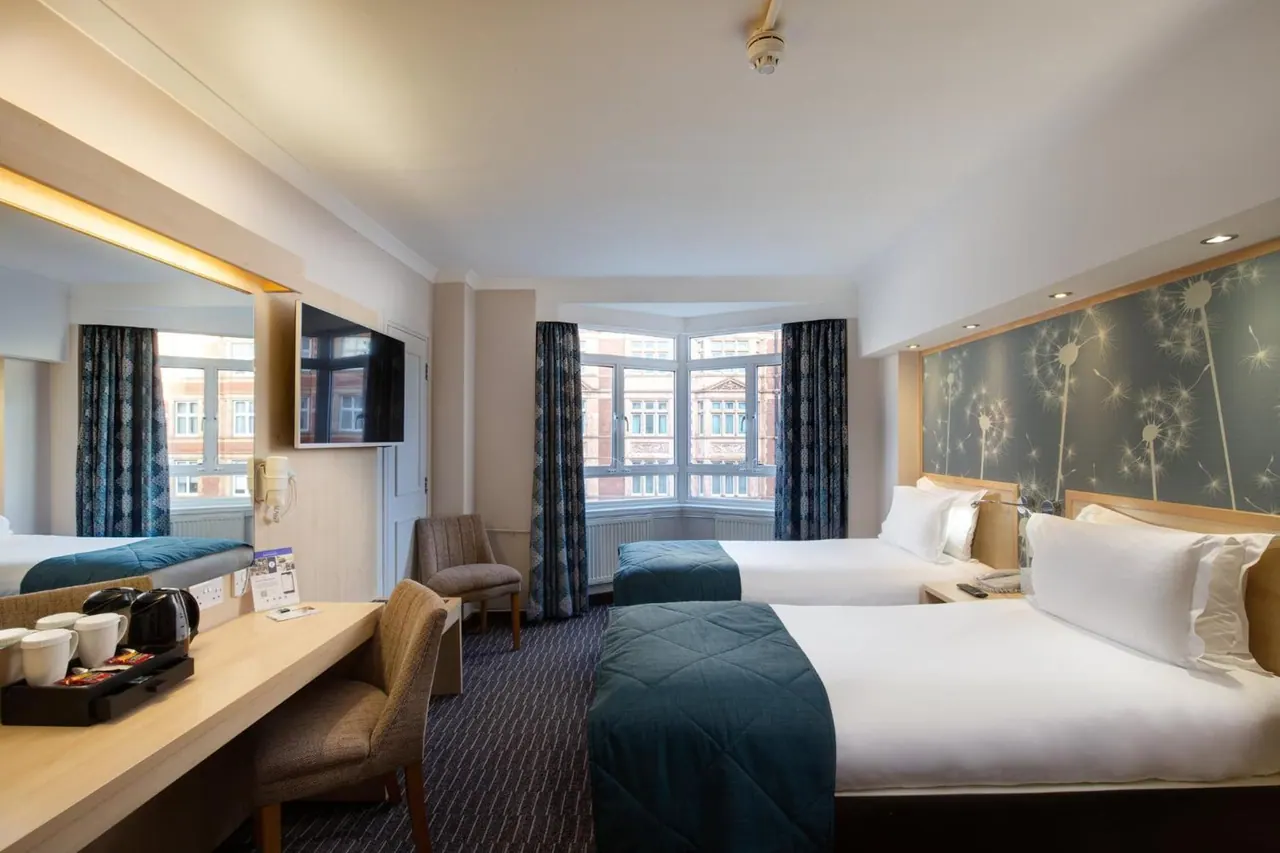 A hotel bedroom with two beds, a wall-mounted television, a desk with a kettle and tea tray, and a bay window looking onto a red-brick building opposite.