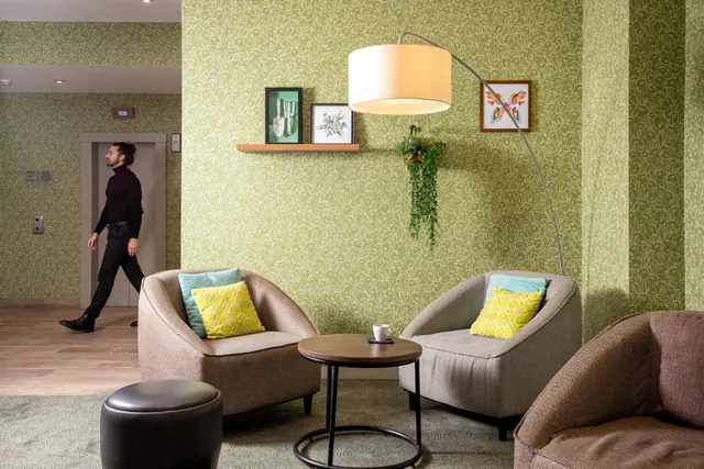 A lobby seating area with two grey armchairs, an arc floor lamp, a round side table and green patterned wallpaper, with a man walking past a lift.