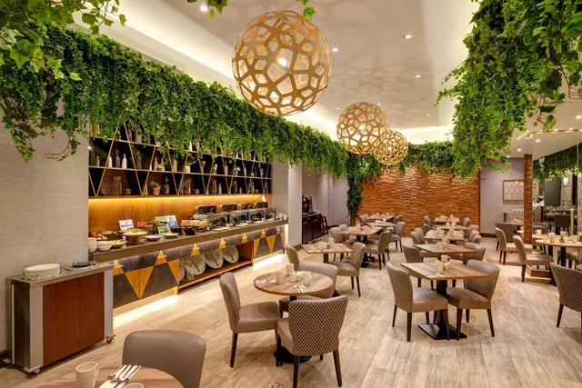 A dining room with upholstered chairs and wooden tables, a buffet counter along one wall, large woven pendant lights and trailing green foliage hanging from the ceiling.