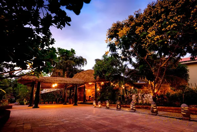 A landscaped hotel entrance courtyard with thatched shelters, tropical trees and illuminated path bollards.
