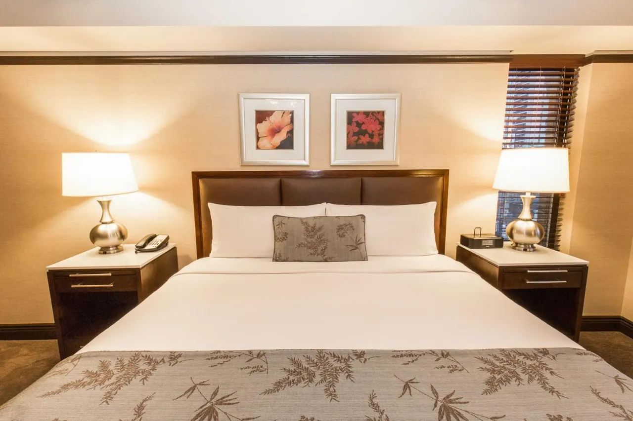 A made king bed with two lit bedside lamps, a telephone and a clock radio on the nightstands, and framed flower prints on the wall above.