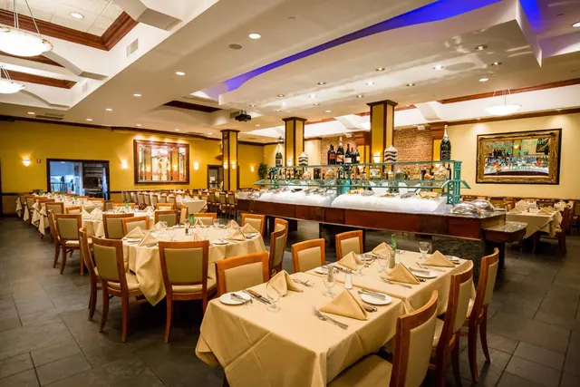 A restaurant dining room with rows of tables set with cream tablecloths and napkins, a food display counter on ice, and a bar with bottles along the back wall.