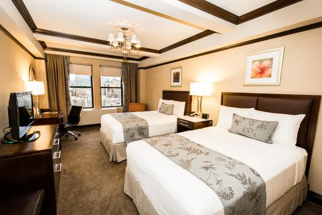 A hotel bedroom with two double beds, a wooden desk with a flat-screen television and office chair, a chandelier, and windows showing city buildings.
