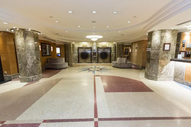An empty hotel lobby with marble columns, elevator doors, wood-panelled walls and a terrazzo floor with a compass star inlay.