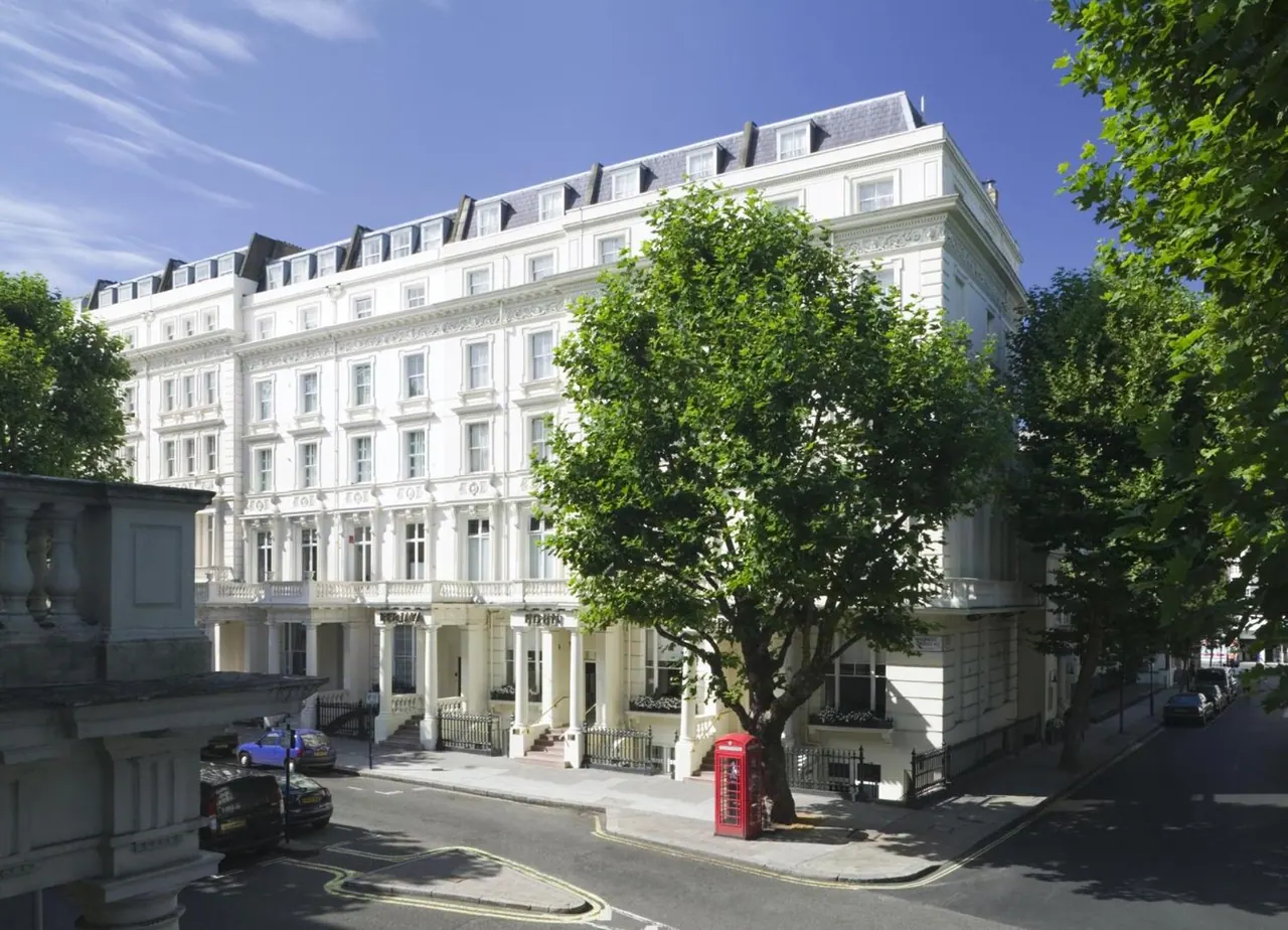 A white stucco hotel building with a large tree in front, a red telephone box on the pavement, and parked cars on the street under a blue sky.