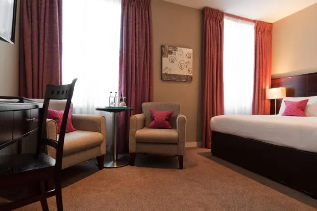 A hotel bedroom with a double bed, two armchairs beside a small round table with water bottles, a desk and a wall-mounted television.