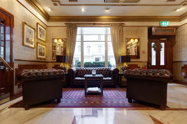 A lobby seating area with two brown leather armchairs and a Chesterfield sofa on a patterned rug, with a large window looking onto a building across the street.