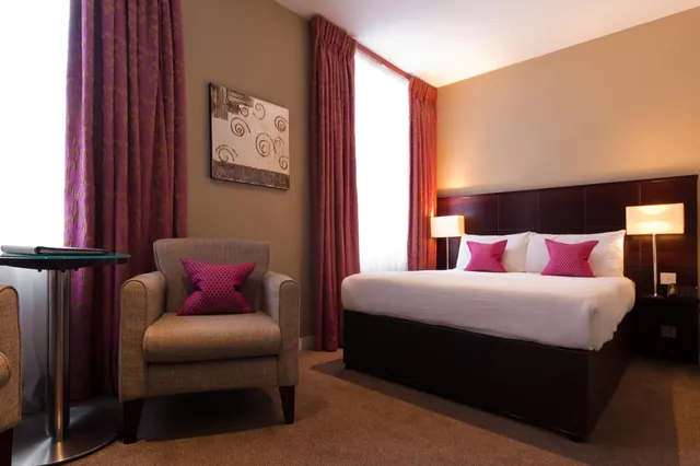 A hotel bedroom with a made double bed with pink cushions, an armchair with a pink cushion, and a small round table beside a curtained window.