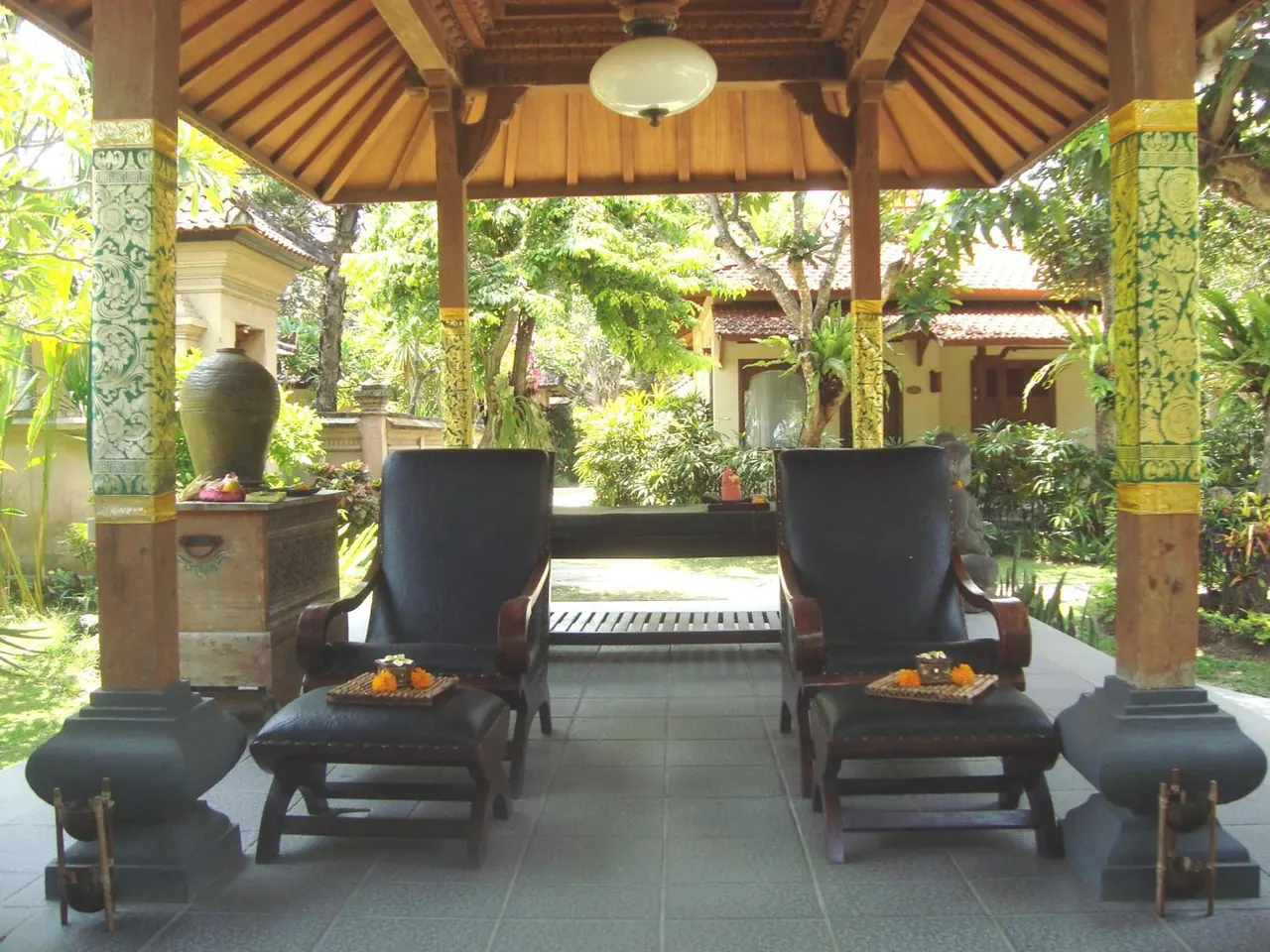 Two leather armchairs with footstools sit under a wooden pavilion roof, facing a garden and a villa building beyond.