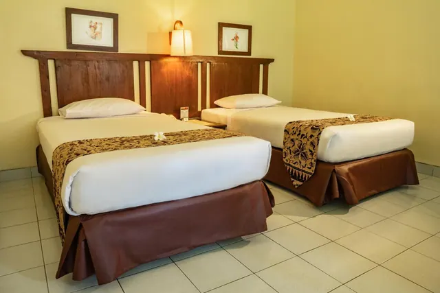 Two single beds with a carved wooden headboard, batik bed runners and framed wall art in a room with cream walls and a tiled floor.