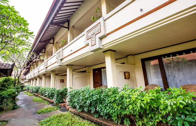Two-storey hotel block with carved cream balconies, wicker chairs behind a hedge on the ground floor, and a paved path lined with trees.