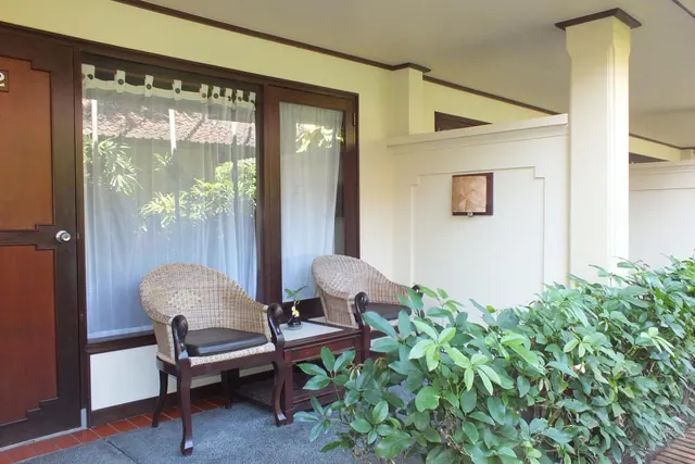 Two wicker armchairs and a small side table sit on a tiled terrace outside a room with a white curtained window, beside a green hedge.