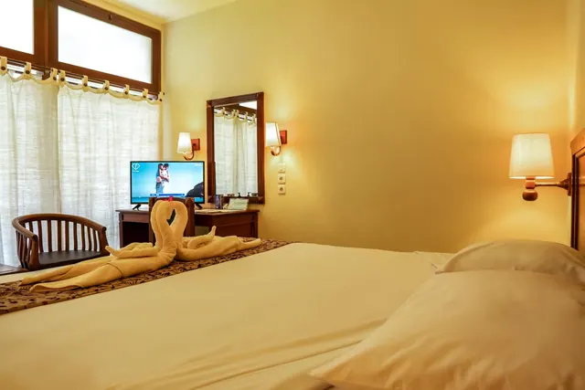 A bedroom with a double bed topped with a folded towel swan, a wooden desk holding a television, a wooden chair, and a window with sheer curtains.