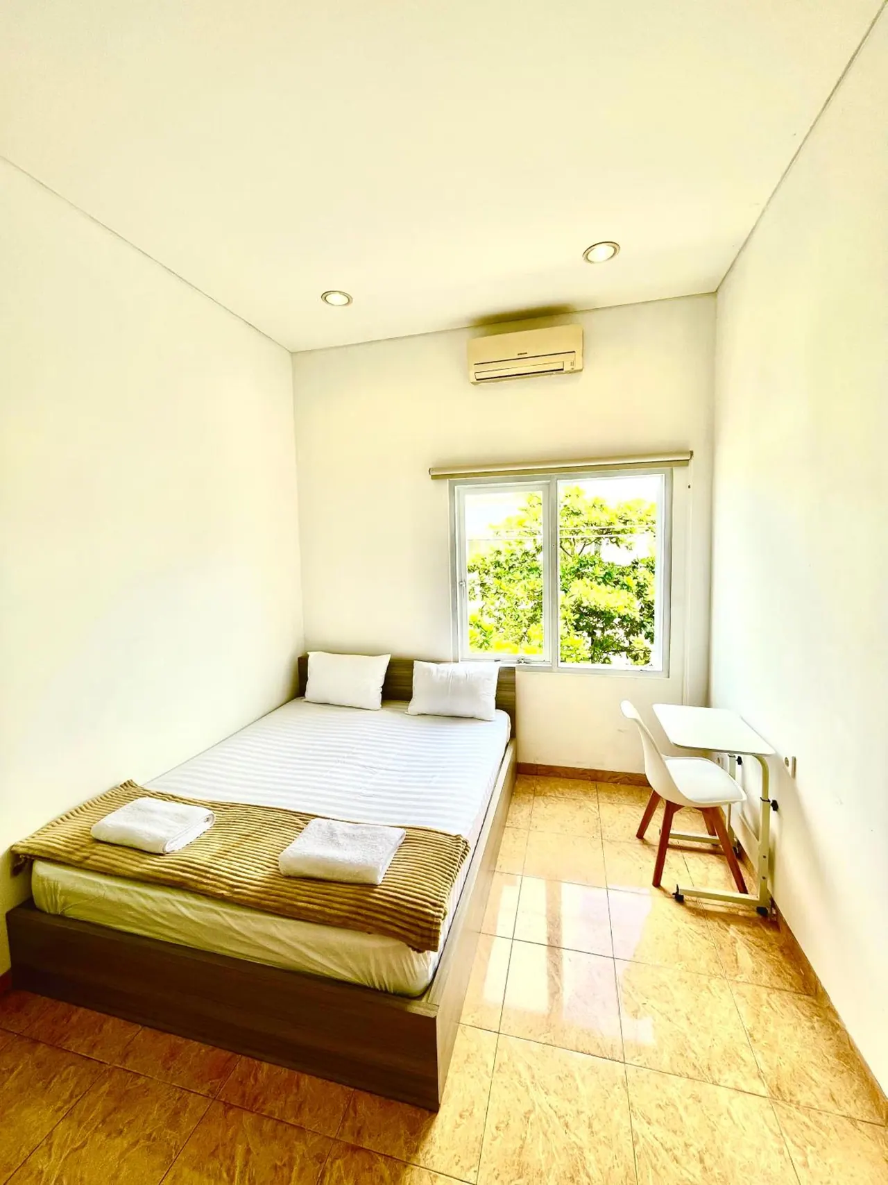 A small bedroom with a double bed, folded towels, a wall-mounted air conditioner, a small desk and chair, and a window showing trees outside.