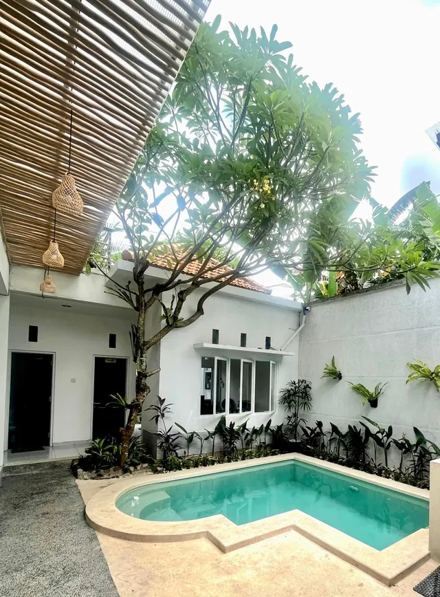 A small pool with a stone surround sits in a courtyard beside a frangipani tree, a white single-storey building and a bamboo overhead screen.