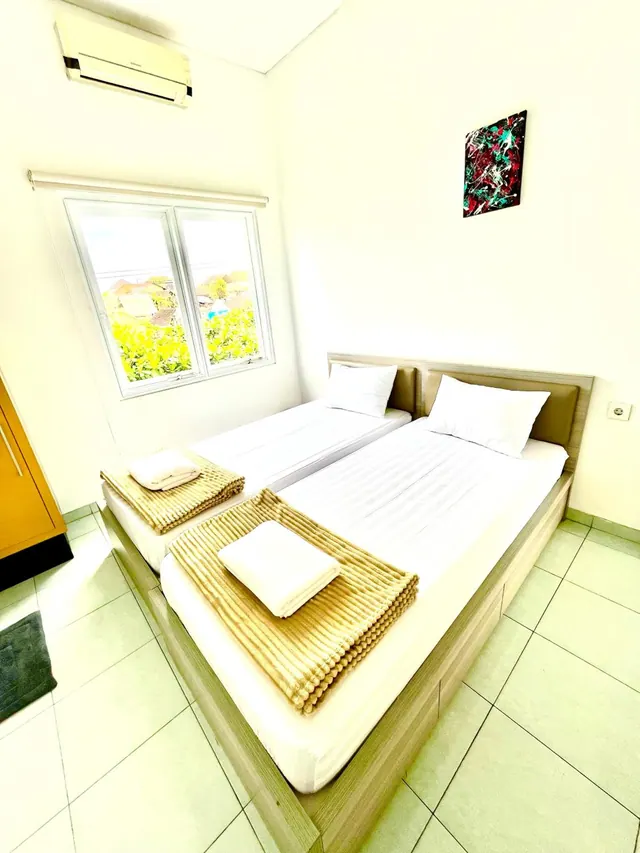 A bedroom with two beds dressed with white linen and folded towels, an air conditioner on the wall, and a window looking out onto yellow-flowering greenery.