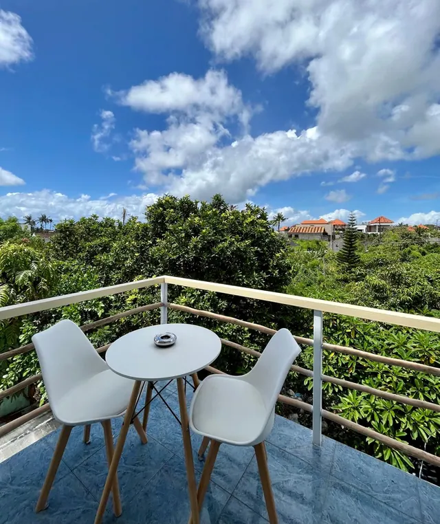 A small round table with two white chairs sits on a tiled balcony, with a railing and a view over trees and rooftops under a blue sky with clouds.