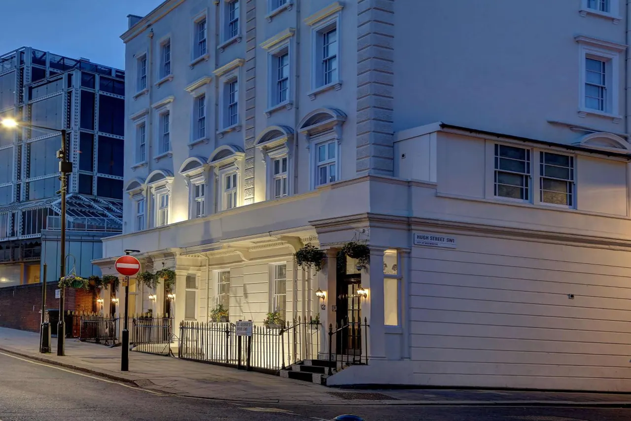 Twilight exterior of a white multi-storey Georgian-style hotel building with lit windows, iron railings and a red no-entry sign on the street corner.