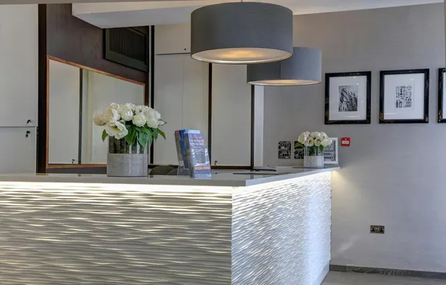 A hotel reception desk with a textured, backlit white front, a vase of white flowers, leaflets, grey pendant lamps and framed black-and-white prints on the wall.