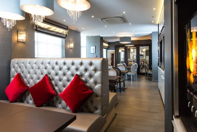 A restaurant dining area with a tufted leather banquette, red cushions, pendant lights, and tables and chairs receding along a wooden floor.