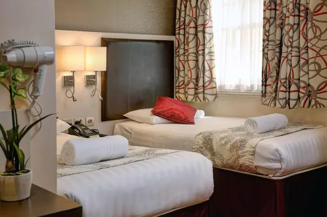 A twin bedroom with two single beds, a dark padded headboard, wall lamps, patterned curtains and a window with sheer net curtains.